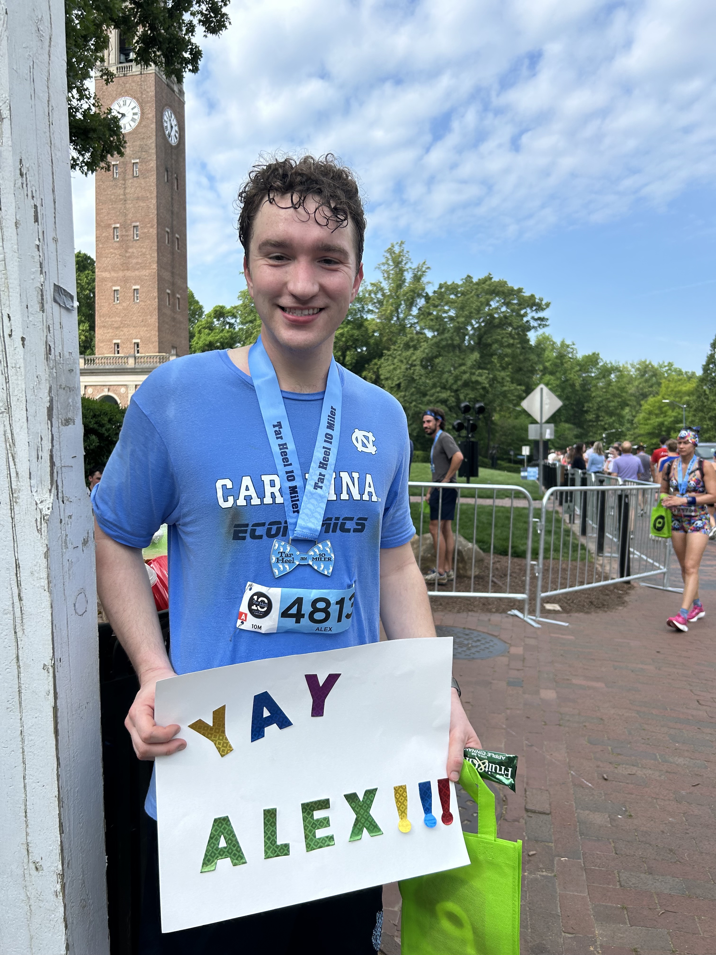 Alex Marsh is smiling in front of the University of North Carolina at Chapel Hill bell tower after completing the 2024 Tar Heel 10 miler. He is very sweat in a Carolina Economics t-shirt and holding a sign that reads "YAY ALEX!!!." He is also wearing the medal for completing the race which is the shape of a bowtie along with his race bib indicating that he was runner 4813.